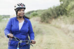 A mature, African American woman riding a mountain bike in a state park. She is wearing a helmet and a blue jacket, looking away from the camera with a confident expression.