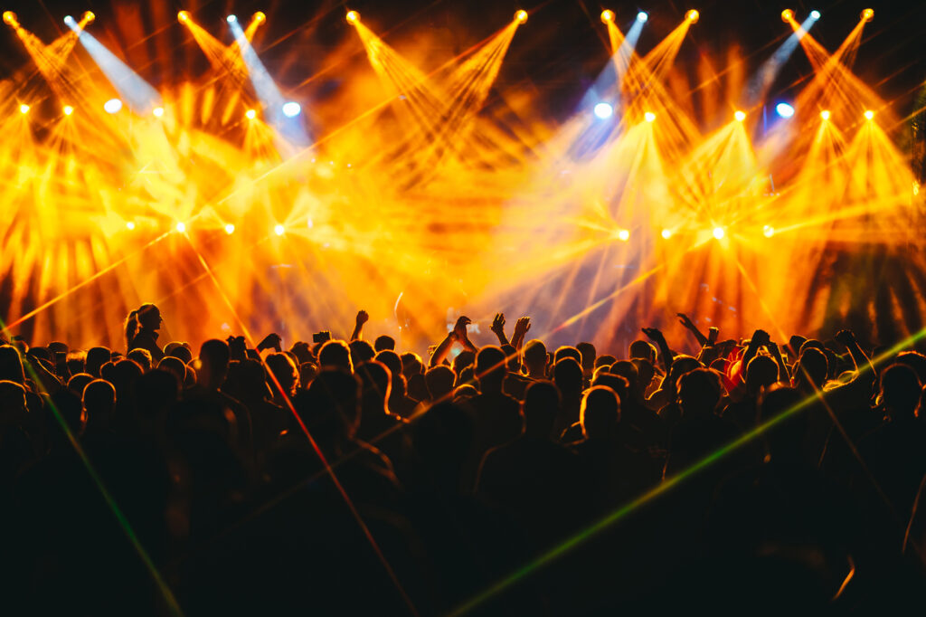 Crowd is enjoying live performance at open air summer festival at night. Nightlife. Silhouettes of people are standing in the dark and listening to electro music at concert. There are orange lights.