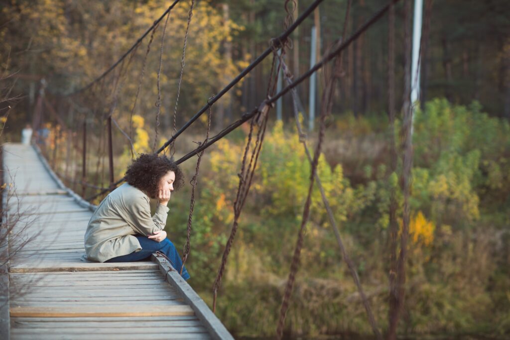 girl with curly hair sits on bridge over river in countryside in autumn. outdoor recreation alone.