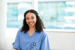 Wearing her scrubs, the female young adult medical student looks at the camera and smiles broadly.