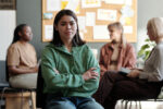 Young brunette woman in green casual pullover and blue jeans looking at camera while sitting against psychologist and patients having session