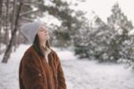 Young beautiful woman wearing fashionable faux fur coat and woolen hat enjoying snowfall in a winter park.